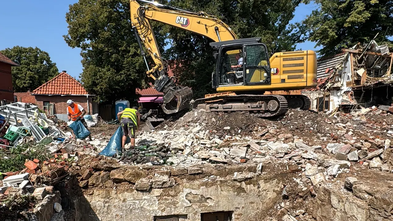 Bagger reißt ein Gebäude ab, davor aufgeschichteter Bauschutt und zwei Arbeiter